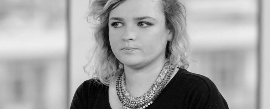 Kobieta z krótkimi, kręconymi włosami w czarnej bluzce i ozdobnym naszyjniku, patrzy w dal.