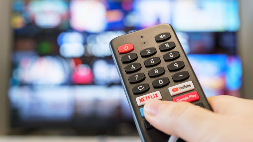 Pilot z przyciskami do TV, trzymany w dłoni, w tle rozmyty ekran telewizora.