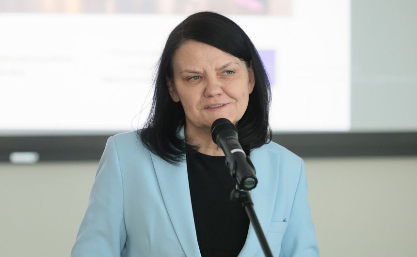 Kobieta z ciemnymi włosami w błękitnej marynarce, mówi do mikrofonu. Tło rozmyte.