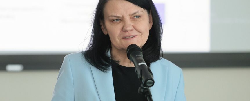 Kobieta z ciemnymi włosami w błękitnej marynarce, mówi do mikrofonu. Tło rozmyte.