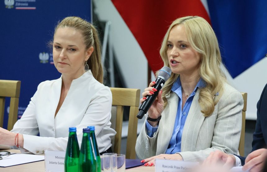 Dwie kobiety podczas spotkania; jedna przemawia, druga słucha, w tle flagi Polski.