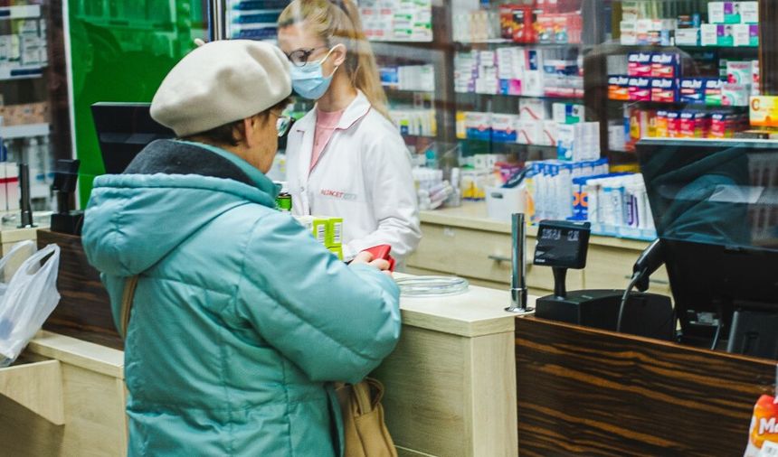 Kobieta w turkusowej kurtce kupuje leki w aptece, pracownica w białym fartuchu obsługuje klientkę.