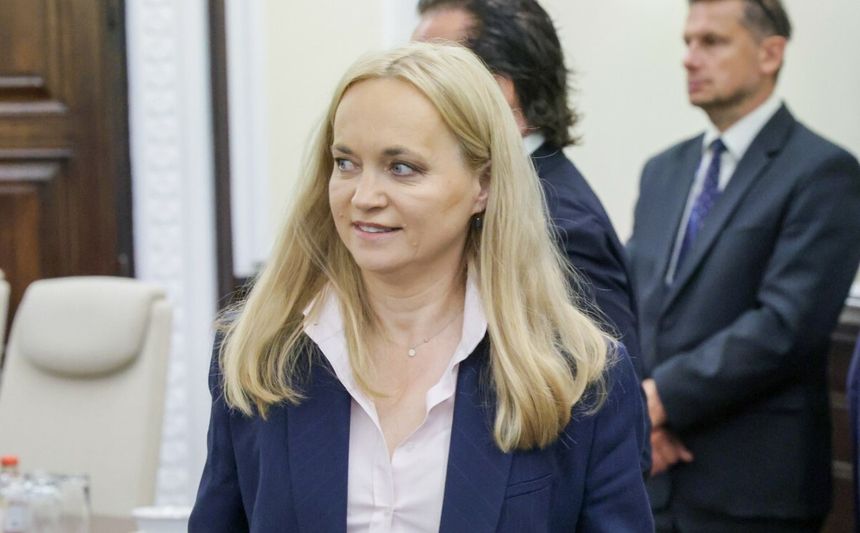 Na zdjęciu uśmiechająca się kobieta w jasnej bluzce i ciemnej marynarce. W tle inni mężczyźni.