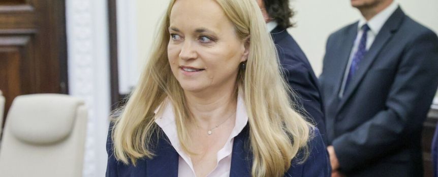 Na zdjęciu uśmiechająca się kobieta w jasnej bluzce i ciemnej marynarce. W tle inni mężczyźni.
