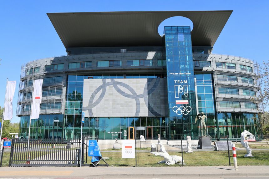 Budynek z dużymi przeszkleniami, logo olimpijskie i rzeźby wokoło. Słoneczny dzień.
