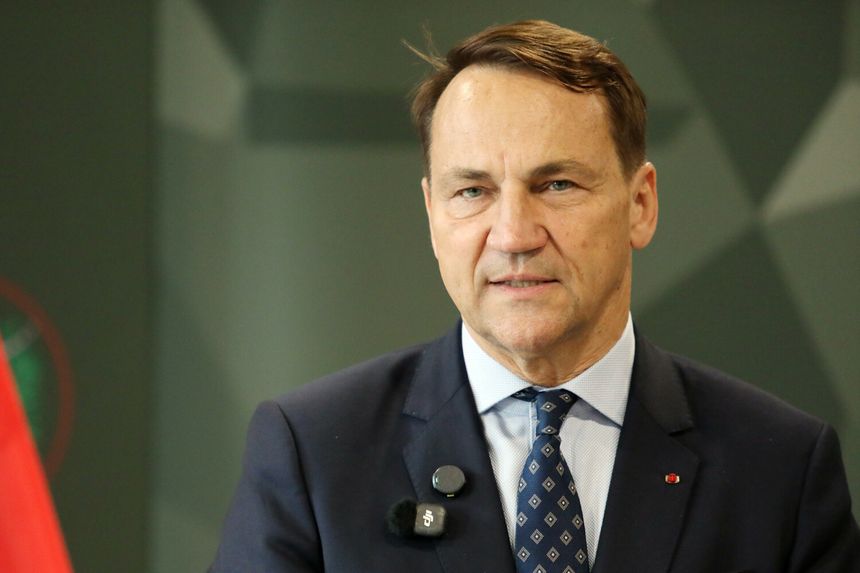 Mężczyzna w garniturze, z mikrofonem, przodem do kamery. Tło w odcieniach zieleni.