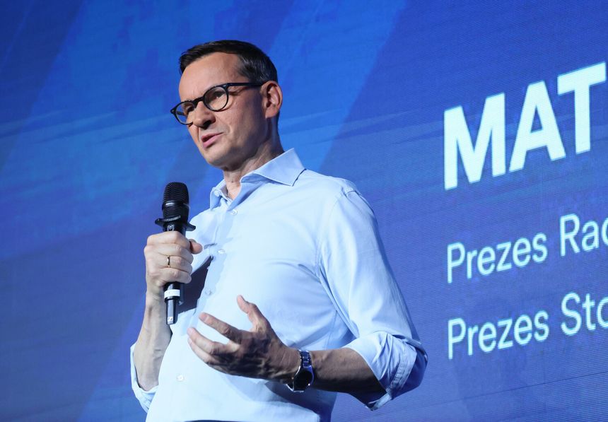 Mężczyzna w okularach, w jasnej koszuli, przemawia do mikrofonu. Na tle niebieskie wzory.