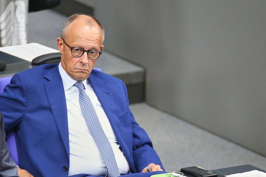 Mężczyzna w niebieskim garniturze siedzi na sali obrad. Obok leży telefon.