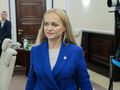 Kobieta w niebieskim żakiecie przechodzi przez pomieszczenie w towarzystwie innych osób.