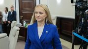 Kobieta w niebieskim żakiecie przechodzi przez pomieszczenie w towarzystwie innych osób.