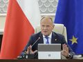 Na zdjęciu Donald Tusk w garniturze, siedzący przy stole z mikrofonem i laptopem. W tle flagi.