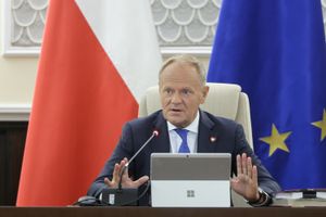 Na zdjęciu Donald Tusk w garniturze, siedzący przy stole z mikrofonem i laptopem. W tle flagi.