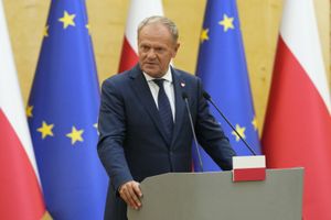 Mężczyzna w garniturze przemawia przy mównicy, w tle flagi Polski i Unii Europejskiej. Donald Tusk.