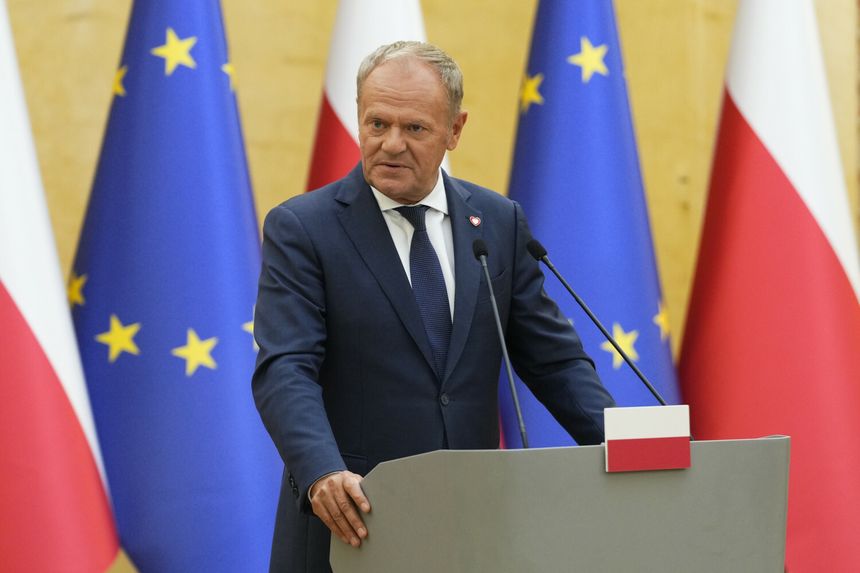 Mężczyzna w garniturze przemawia przy mównicy, w tle flagi Polski i Unii Europejskiej. Donald Tusk.
