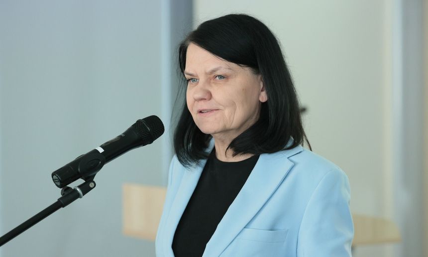 Kobieta w jasnoniebieskim żakiecie, mówi do mikrofonu w tle neutralne ściany.