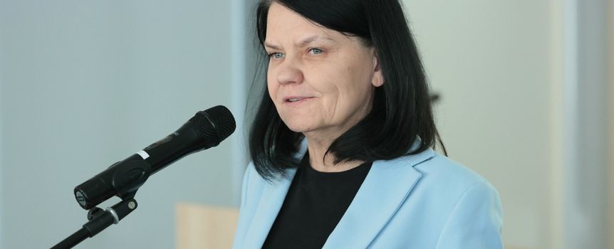 Kobieta w jasnoniebieskim żakiecie, mówi do mikrofonu w tle neutralne ściany.