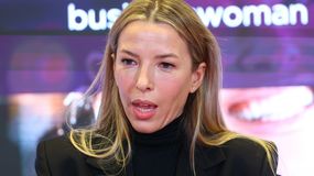 Ewa Chodakowska z długimi, jasnymi włosami w czarnym ubraniu, mówi do kamery na tle rozmytego ekranu.