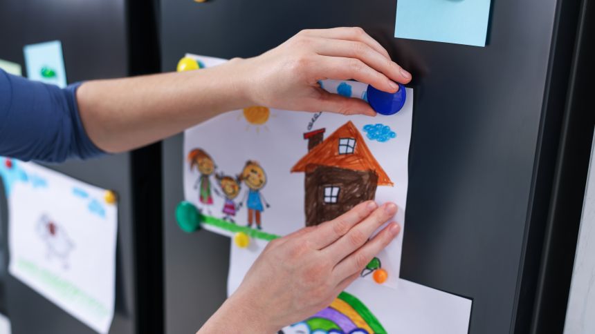 Osoba przyczepia kolorowy rysunek przedstawiający dom do lodówki za pomocą magnesu.