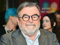 Mężczyzna z okularami i siwą brodą, siedzi w tłumie, uśmiechając się do kamery.