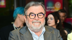 Mężczyzna z okularami i siwą brodą, siedzi w tłumie, uśmiechając się do kamery.