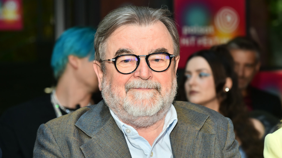 Mężczyzna z okularami i siwą brodą, siedzi w tłumie, uśmiechając się do kamery.