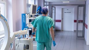 Osoba w zielonym stroju medycznym idzie korytarzem szpitalnym, w tle urządzenia medyczne.