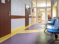 Korytarz w budynku, z drzwiami i niebieskimi oraz białymi krzesłami. Osób brak.