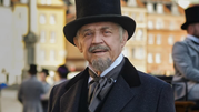 Mężczyzna w kapeluszu cylindrycznym i eleganckim płaszczu, uśmiechający się na tle miasta.