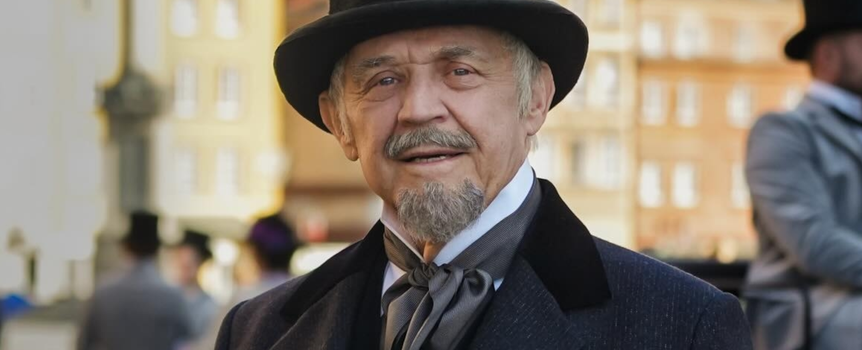 Mężczyzna w kapeluszu cylindrycznym i eleganckim płaszczu, uśmiechający się na tle miasta.