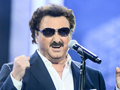 Mężczyzna w ciemnych okularach przeciwsłonecznych śpiewa do mikrofonu na scenie.