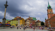 Rynek w Warszawie z kolorowymi budynkami, monumentem i turystami w chmurzystym niebie.