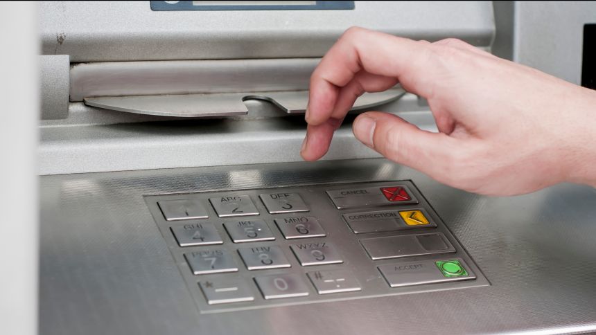 Ręka nad klawiaturą bankomatu, gotowa do nacisnięcia klawisza. Metalowa obudowa urządzenia.