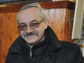 Mężczyzna z uśmiechem, w okularach, w czarnej kurtce, na tle drewnianej ściany.
