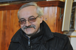 Mężczyzna z uśmiechem, w okularach, w czarnej kurtce, na tle drewnianej ściany.