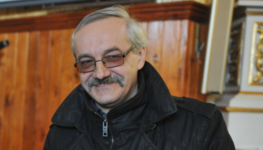 Mężczyzna z uśmiechem, w okularach, w czarnej kurtce, na tle drewnianej ściany.