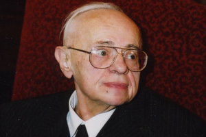 Na zdjęciu mężczyzna z okularami, w eleganckim garniturze, siedzi na tle czerwonej tapety.