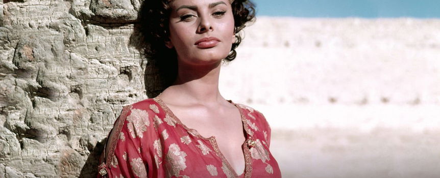 Sophia Loren. Na zdjęciu kobieta o ciemnych włosach w koronkowej bluzce, stojąca przy kamiennej ścianie.