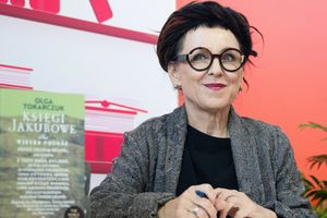 Na zdjęciu widoczna kobieta w okularach, uśmiechająca się, trzymająca długopis, książki w tle.