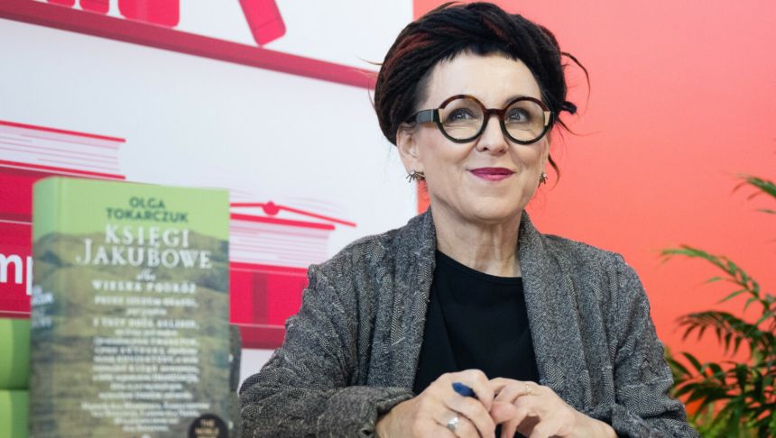Na zdjęciu widoczna kobieta w okularach, uśmiechająca się, trzymająca długopis, książki w tle.