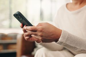Osoba trzymająca telefon w dłoniach, siedząca w jasnym wnętrzu, ubrana w jasny sweter.