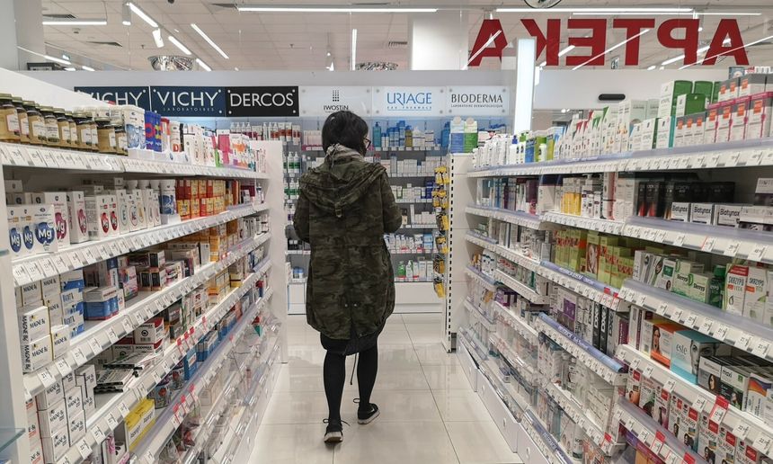 Osoba w kurtce moro stoi w aptece, przeglądając półki z lekami i kosmetykami.