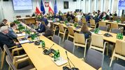 Spotkanie w sali, uczestnicy przy stołach, w tle flagi Polski i UE. Szklane butelki na stołach.