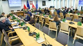 Spotkanie w sali, uczestnicy przy stołach, w tle flagi Polski i UE. Szklane butelki na stołach.