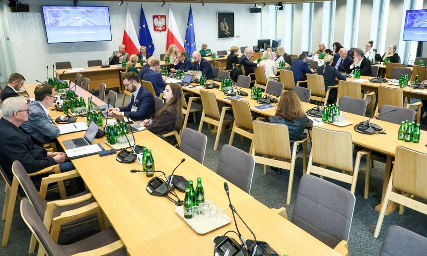 Spotkanie w sali, uczestnicy przy stołach, w tle flagi Polski i UE. Szklane butelki na stołach.