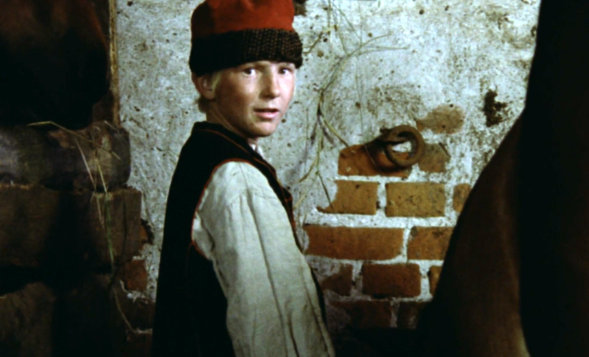 Chłopiec w tradycyjnym ubraniu stoi w stajni obok koni, spoglądając w stronę kamery.