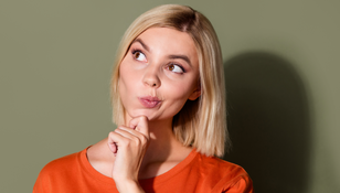Kobieta z krótkimi blond włosami, w pomarańczowej koszulce, zamyślona, dotyka podbródka.