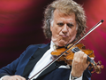 Andre Rieu. Mężczyzna gra na skrzypcach, ma długie, kręcone włosy i elegancki garnitur. Tło czerwone.
