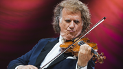 Andre Rieu. Mężczyzna gra na skrzypcach, ma długie, kręcone włosy i elegancki garnitur. Tło czerwone.