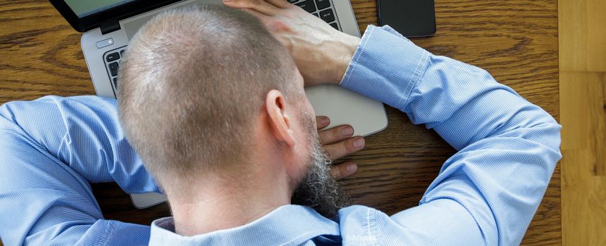 Mężczyzna w niebieskiej koszuli siedzi przy laptopie, opierając się na nim z rękami na klawiaturze.