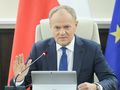 Mężczyzna w garniturze siedzi przy stole, gestykulując, z flagami w tle. Donald Tusk.